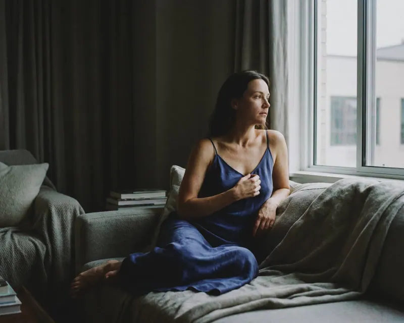 Mujer sentada en sillón azul mirando por la ventana, sesión boudoir íntima luz tarde