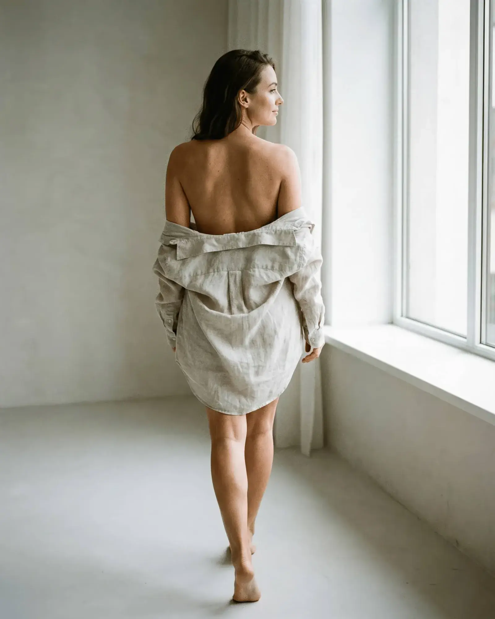 Mujer de espaldas junto a ventana con camisa caída y paredes blancas, sesión boudoir