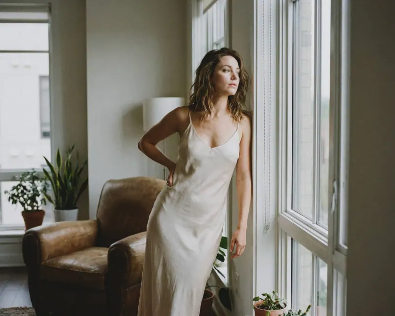 Mujer con vestido blanco de seda con mirada lateral y luz suave junto a plantas, boudoir
