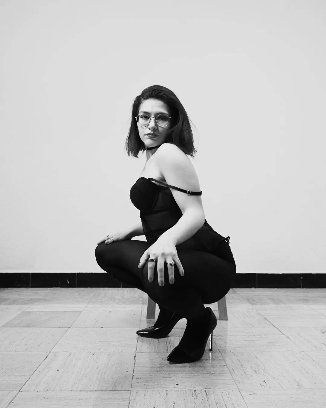 Mujer en cuclillas con corsé negro y mirada directa, boudoir de estudio en blanco y negro