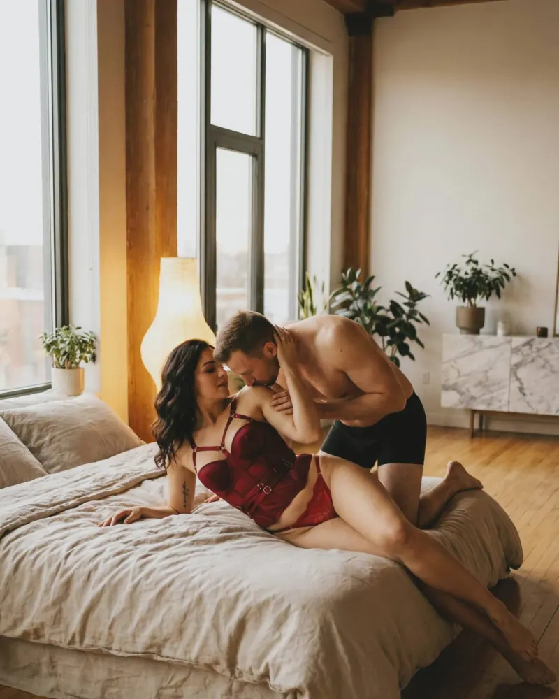 Pareja boudoir en cama de loft con beso en el hombro y corsé rojo