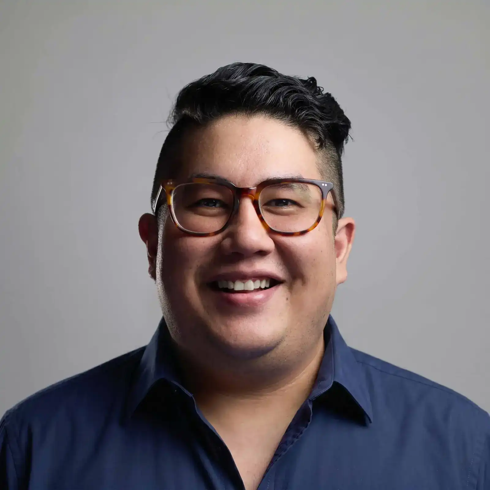 Retrato de Thoru García, fotógrafo boudoir en CDMX, con lentes de pasta, sonriendo y camisa azul, sobre un fondo gris.